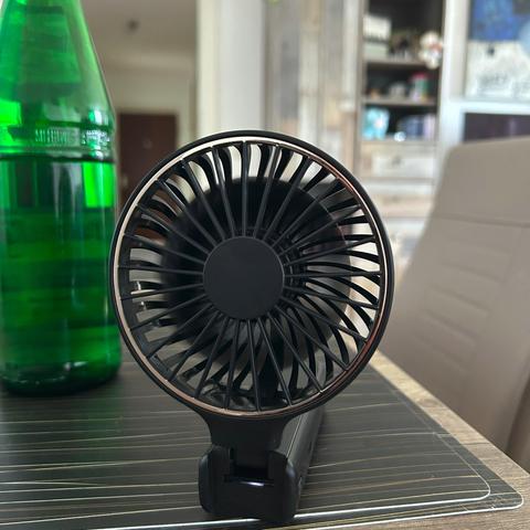 Ein kleiner schwarzer Ventilator mit Kupferrand steht neben einer grünen Glasflasche auf einem dunklen Tisch. Im Hintergrund ist ein Regal mit verschiedenen Gegenständen zu sehen.