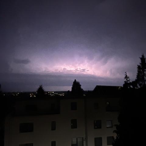 Ein Gewitter bei Nacht mit Blitzen am Himmel über einer Stadt und einigen Gebäuden im Vordergrund.