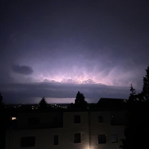 Ein Gewitter bei Nacht mit Blitzen am Himmel über einer Stadt und einigen Gebäuden im Vordergrund.