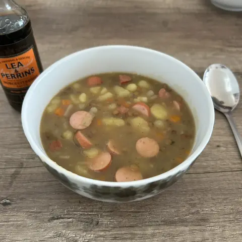 Eine Schüssel Linsensuppe mit Würstchenscheiben, Kartoffeln und Karotten, daneben eine Flasche Worcestershire-Sauce und ein Löffel.